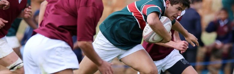 Nahaufnahme eines Rugby-Spielers im grünen Trikot mit roten und weißen Streifen, der den ovalen Rugbyball fest im Arm hält und vorwärts rennt. Er wird eng von Gegenspielern in weinroten Trikots bedrängt.