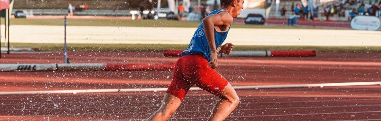 Seitliche Aufnahme eines Leichtathleten in blauem Oberteil und roter Laufhose, der auf einer roten Laufbahn sprintet. Um seine Beine herum spritzt Wasser auf, das im Sonnenlicht glänzt.